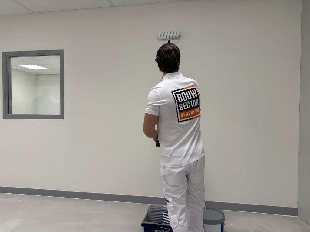 Schilder in witte werkkleding rolt een lichte wand in een lege ruimte, verfbak op de vloer