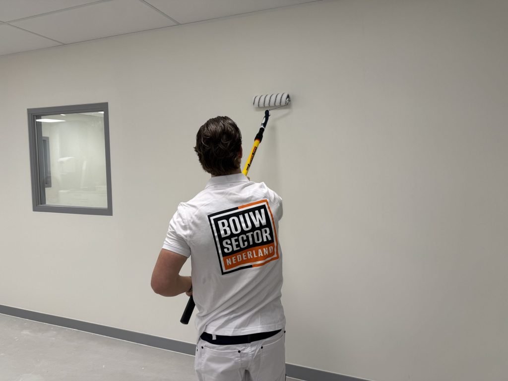 Schilder met Bouwsector-logo op de rug rolt een witte wand in een lege ruimte met binnenraam