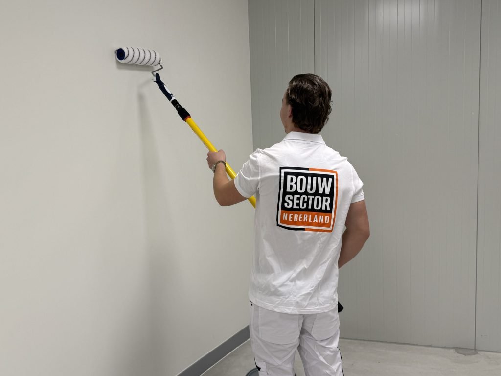 Schilder in wit poloshirt met Bouwsector-logo zet de roller hoog op een lichte muur