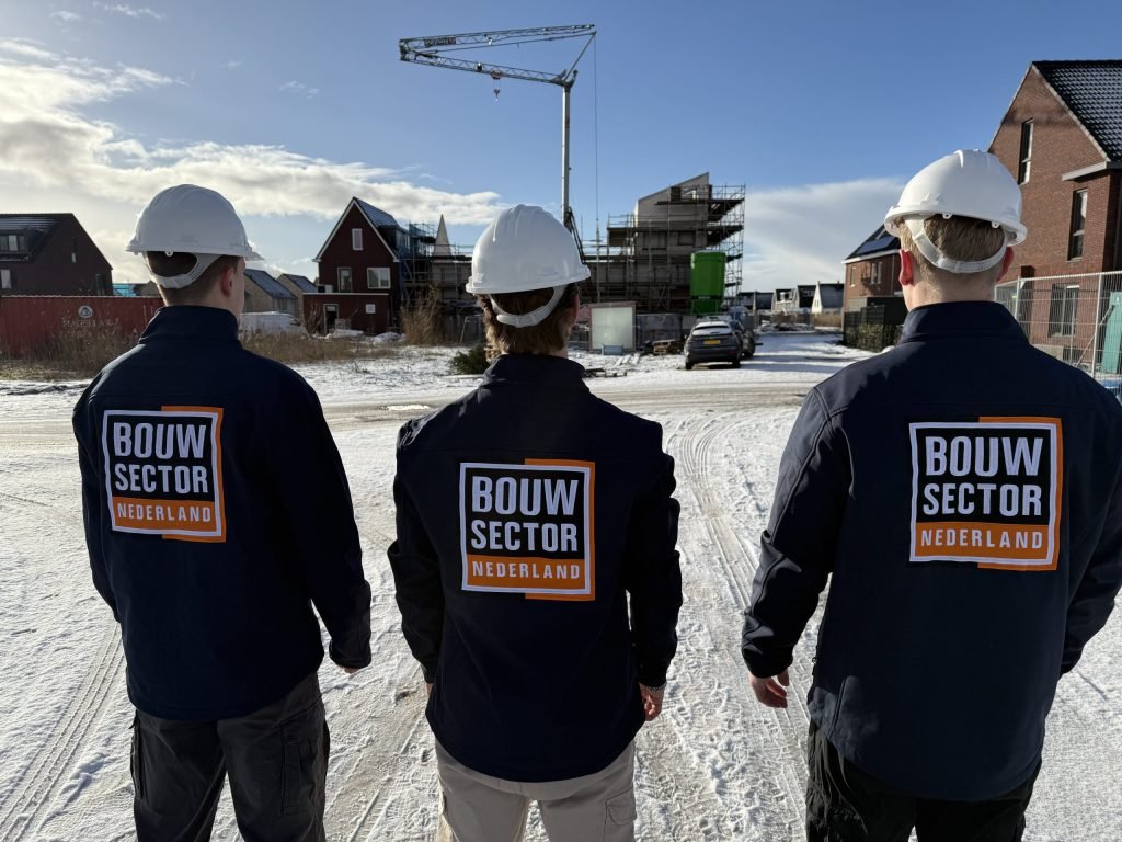 Drie vakmensen van Bouwsector Nederland met witte bouwhelm lopen in de sneeuw naar een rij bakstenen woningen