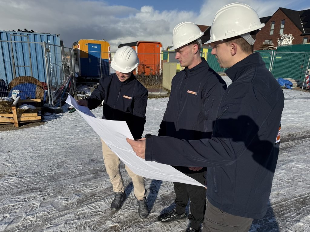 Drie vakmensen van Bouwsector Nederland met witte helm bekijken een bouwtekening op een besneeuwd bouwterrein